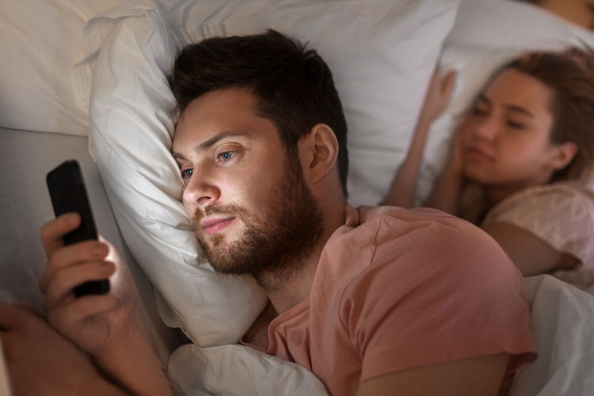 un homme en couple sur son téléphone au lit avec sa femme parlant à une autre femme