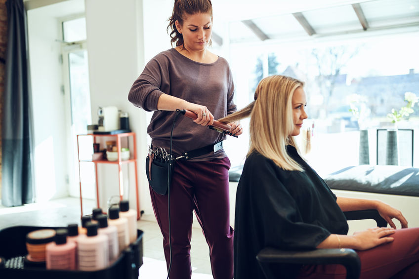 Une femme blonde se fait lisser les cheveux avec un fer par une coiffeuse dans un salon de coiffure